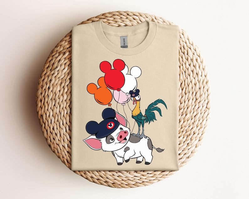 Op de afbeelding: Beige T-shirt met een cartoonvarken met een marineblauwe hoed met een Mickey Mouse-ontwerp. Een haan en kleurrijke ballonnen zijn ook aanwezig. Het shirt ligt op een geweven, cirkelvormig oppervlak.