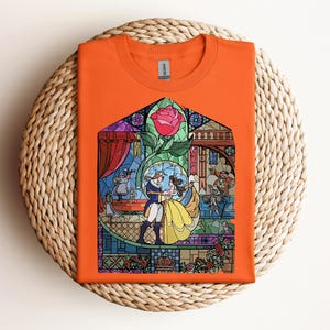 May include: Orange t-shirt featuring a stained-glass design with characters from a fairy tale. The design includes a rose, a couple dancing, and other characters. The shirt is folded on a woven, circular surface.