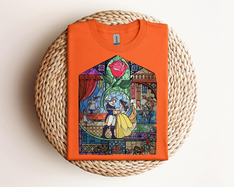 May include: Orange t-shirt featuring a stained-glass design with characters from a fairy tale. The design includes a rose, a couple dancing, and other characters. The shirt is folded on a woven, circular surface.