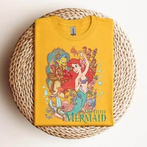 May include: Mustard yellow t-shirt featuring a colorful graphic of Ariel from The Little Mermaid. The design includes Ariel, Flounder, and other sea creatures, with the words "The Little Mermaid" in a teal font. The shirt is folded on a woven surface.