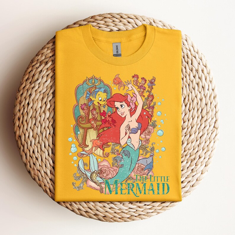 May include: Mustard yellow t-shirt featuring a colorful graphic of Ariel from The Little Mermaid. The design includes Ariel, Flounder, and other sea creatures, with the words "The Little Mermaid" in a teal font. The shirt is folded on a woven surface.