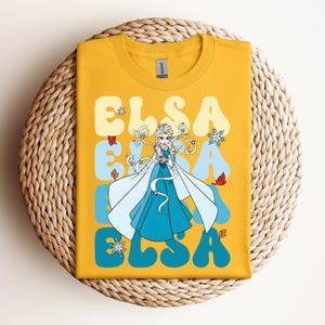 Op de afbeelding: Mosterdgeel T-shirt met een afbeelding van Elsa uit Frozen. Het ontwerp bevat de naam "ELSA" in een retro lettertype, met een cartoonafbeelding van Elsa in een blauwe jurk en een witte cape. Het shirt wordt weergegeven op een geweven, cirkelvormig oppervlak.