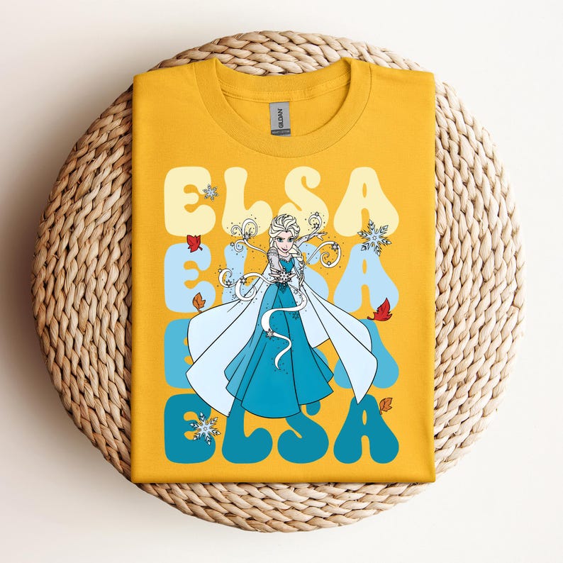 Op de afbeelding: Mosterdgeel T-shirt met een afbeelding van Elsa uit Frozen. Het ontwerp bevat de naam "ELSA" in een retro lettertype, met een cartoonafbeelding van Elsa in een blauwe jurk en een witte cape. Het shirt wordt weergegeven op een geweven, cirkelvormig oppervlak.