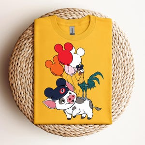 Op de afbeelding: Mosterdgeel t-shirt met een cartoonillustratie van een varken met een hoed, een haan en ballonnen. Het varken is wit met zwarte vlekken en een roze neus. De ballonnen zijn rood, oranje en wit. De haan is blauw en groen.