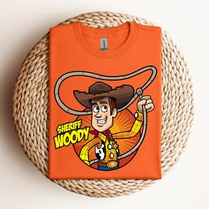 May include: Orange t-shirt featuring a cartoon illustration of Sheriff Woody from Toy Story. The design includes Woody's hat, rope, and the text "Sheriff Woody" in yellow and green. The shirt is folded on a woven surface.