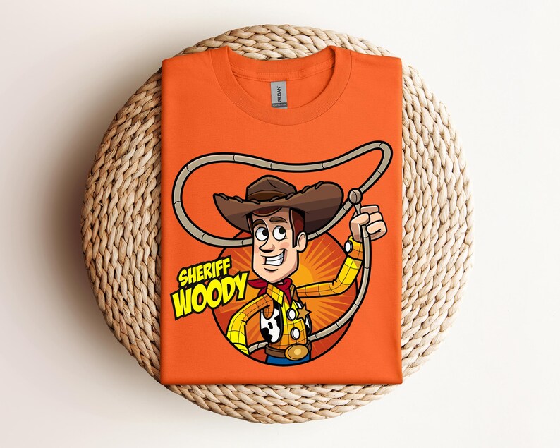 May include: Orange t-shirt featuring a cartoon illustration of Sheriff Woody from Toy Story. The design includes Woody's hat, rope, and the text "Sheriff Woody" in yellow and green. The shirt is folded on a woven surface.