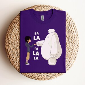 K&ouml;nnte beinhalten: Lilafarbenes T-Shirt mit einem wei&szlig;en Cartoon-Roboter und einer Jungenfigur. Der Text "BA LA LA LA" ist auf dem T-Shirt aufgedruckt.