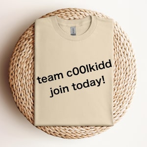 May include: A beige crew neck t-shirt with the text "team c00lkidd join today!" in black. The shirt is displayed on a round, woven, beige placemat.