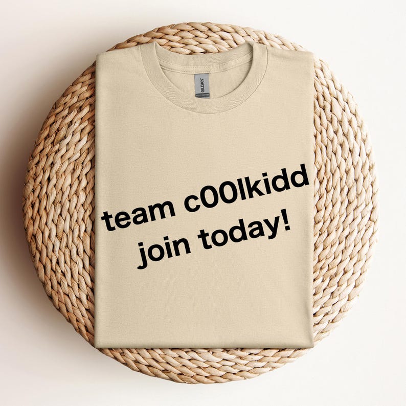 May include: A beige crew neck t-shirt with the text "team c00lkidd join today!" in black. The shirt is displayed on a round, woven, beige placemat.