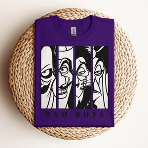 May include: A purple t-shirt featuring a graphic design of four cartoon villains in black and white, each in a vertical panel. The text "BAD BOYS" is printed in black at the bottom of the design. The shirt is folded on a woven, circular surface.