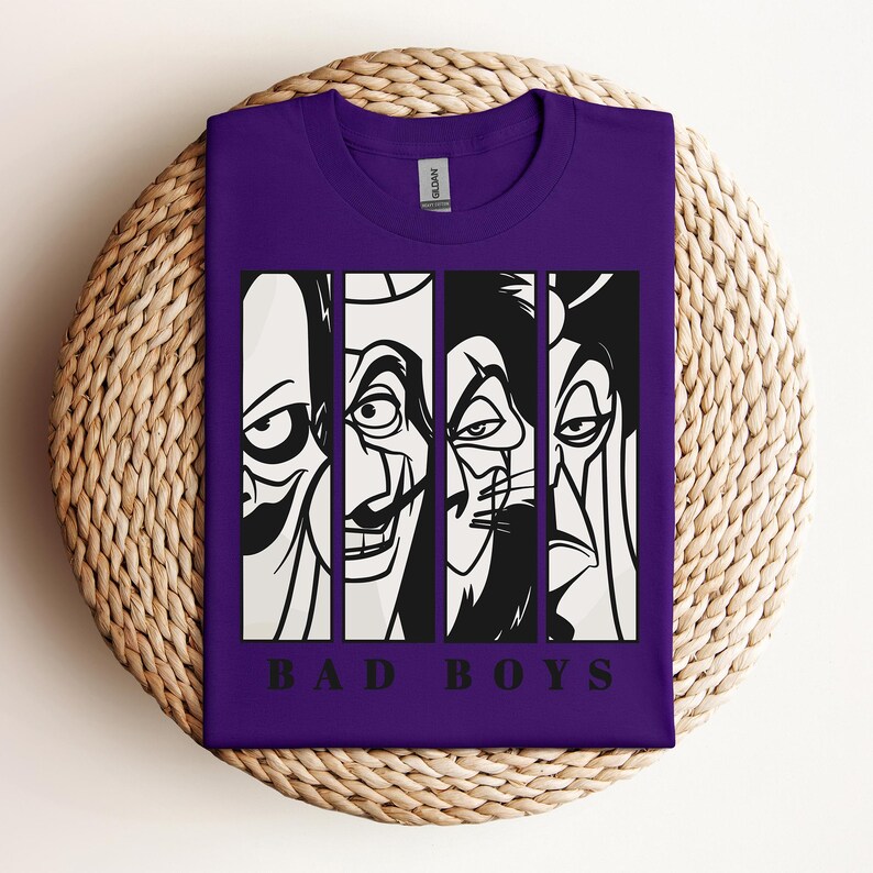 May include: A purple t-shirt featuring a graphic design of four cartoon villains in black and white, each in a vertical panel. The text "BAD BOYS" is printed in black at the bottom of the design. The shirt is folded on a woven, circular surface.