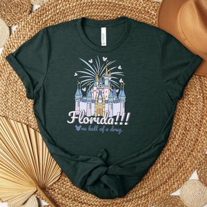 Puede incluir: Camiseta verde oscuro con una ilustraci&oacute;n de castillo, fuegos artificiales y el texto "Florida!!! no hell of a drug." El dise&ntilde;o incorpora siluetas de Mickey Mouse. La camiseta est&aacute; hecha de un material suave.