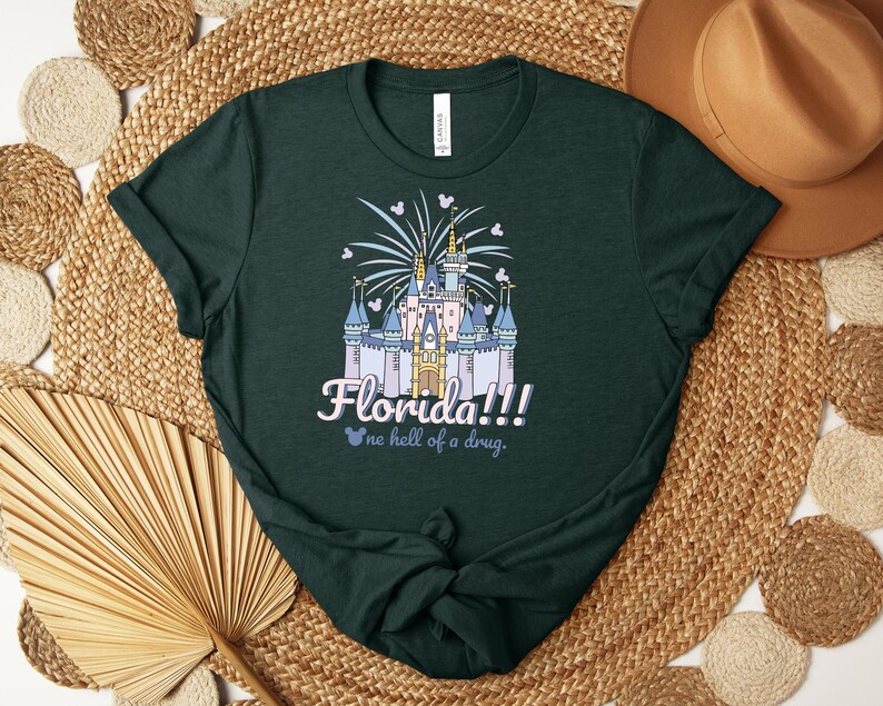 Puede incluir: Camiseta verde oscuro con una ilustraci&oacute;n de castillo, fuegos artificiales y el texto "Florida!!! no hell of a drug." El dise&ntilde;o incorpora siluetas de Mickey Mouse. La camiseta est&aacute; hecha de un material suave.