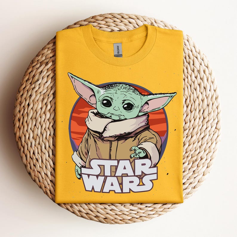 Op de afbeelding: Mosterdgeel T-shirt met een afbeelding van een groen alien-personage met grote oren, tegen een rode en oranje achtergrond. De woorden "STAR WARS" zijn in het wit onder het personage gedrukt. Het shirt wordt gepresenteerd op een geweven, cirkelvormig oppervlak.