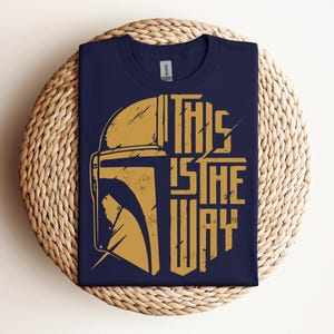 May include: Navy blue t-shirt with a gold graphic of a helmet and the text "THIS IS THE WAY". The design is distressed, giving it a vintage look. The shirt is folded on a woven, circular surface.