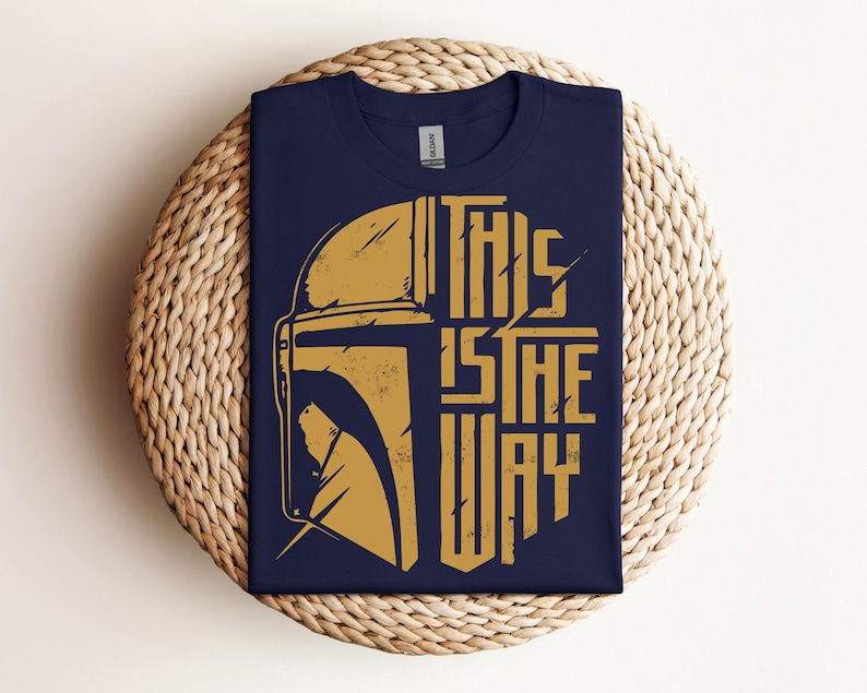 May include: Navy blue t-shirt with a gold graphic of a helmet and the text "THIS IS THE WAY". The design is distressed, giving it a vintage look. The shirt is folded on a woven, circular surface.