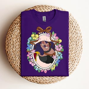 Puede incluir: Una camiseta morada con un personaje de dibujos animados dentro de un marco ovalado. El marco est&aacute; decorado con un lazo marr&oacute;n, flores coloridas y un borde de perlas. El personaje es azul oscuro y marr&oacute;n.