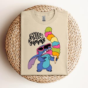 May include: Beige t-shirt featuring a cartoon character wearing sunglasses and holding a tall, colorful ice cream cone. The words "hello summer" are written in a playful font above the character.