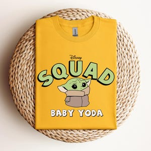 May include: Mustard yellow t-shirt featuring the text "Disney Squad" above an illustration of Baby Yoda. The words "Baby Yoda" are printed below the image. The shirt is folded and placed on a woven, circular surface.