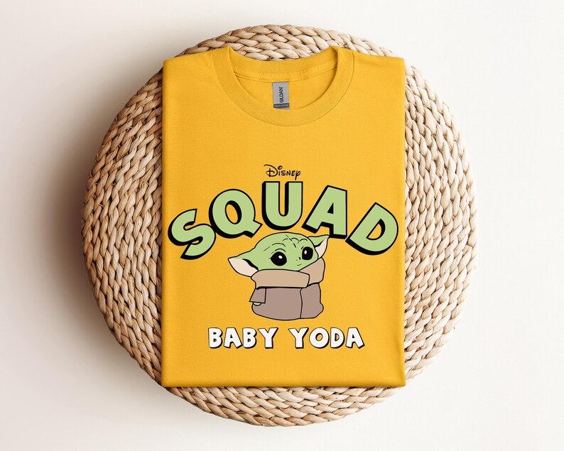 May include: Mustard yellow t-shirt featuring the text "Disney Squad" above an illustration of Baby Yoda. The words "Baby Yoda" are printed below the image. The shirt is folded and placed on a woven, circular surface.