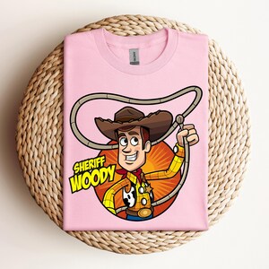 May include: A light pink t-shirt featuring a cartoon illustration of Sheriff Woody from Toy Story. The graphic includes Woody in his cowboy attire, holding a lasso, with the words "Sheriff Woody" in a yellow and green font.