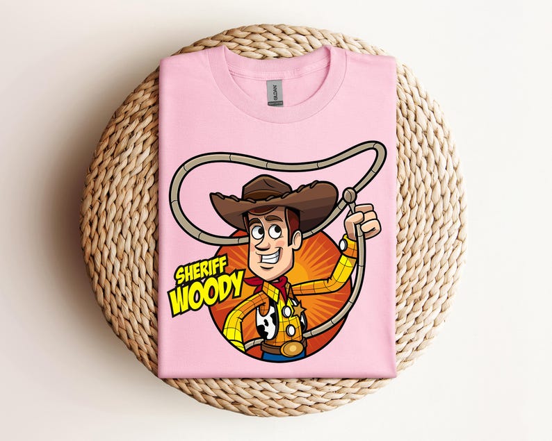 May include: A light pink t-shirt featuring a cartoon illustration of Sheriff Woody from Toy Story. The graphic includes Woody in his cowboy attire, holding a lasso, with the words "Sheriff Woody" in a yellow and green font.