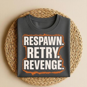 Può includere: T-shirt grigio scuro con le parole "RESPAWN. RETRY. REVENGE." in bianco, bordate di arancione. La maglietta è esposta su un sottobicchiere rotondo e intrecciato beige.