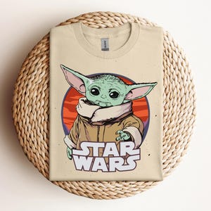 Op de afbeelding: Beige T-shirt met een afbeelding van een groen alien-personage met grote oren, tegen een rode en oranje cirkelvormige achtergrond. De woorden "STAR WARS" zijn in het wit onder het personage gedrukt. Het shirt wordt gepresenteerd op een geweven oppervlak.