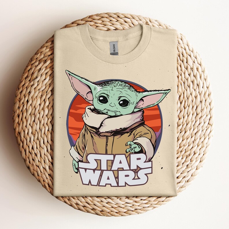 Op de afbeelding: Beige T-shirt met een afbeelding van een groen alien-personage met grote oren, tegen een rode en oranje cirkelvormige achtergrond. De woorden "STAR WARS" zijn in het wit onder het personage gedrukt. Het shirt wordt gepresenteerd op een geweven oppervlak.