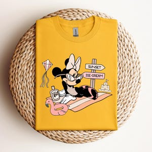 K&ouml;nnte beinhalten: Gelbes T-Shirt mit einem Cartoon-Bild von Minnie Mouse, die eine Sonnenbrille und eine rosa Schleife tr&auml;gt. Sie liegt auf einem Strandtuch mit einem rosa Flamingo-Schwimmring, einer Flasche Sonnencreme und einem Schild mit der Aufschrift "Sunset" und "Ice-Cream".