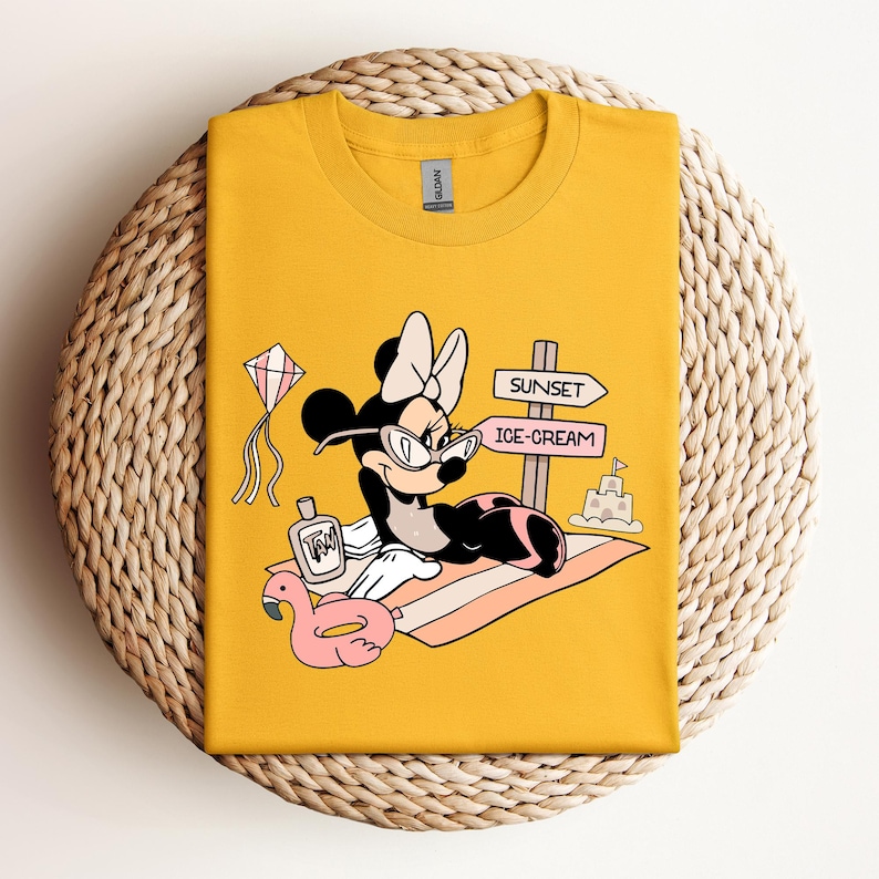 K&ouml;nnte beinhalten: Gelbes T-Shirt mit einem Cartoon-Bild von Minnie Mouse, die eine Sonnenbrille und eine rosa Schleife tr&auml;gt. Sie liegt auf einem Strandtuch mit einem rosa Flamingo-Schwimmring, einer Flasche Sonnencreme und einem Schild mit der Aufschrift "Sunset" und "Ice-Cream".