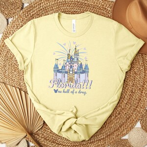 Puede incluir: Camiseta amarilla clara con una ilustraci&oacute;n colorida de un castillo. El texto dice "Florida!!! one hell of a drug." El dise&ntilde;o del castillo incluye detalles en azul, morado y dorado, con fuegos artificiales y corazones.