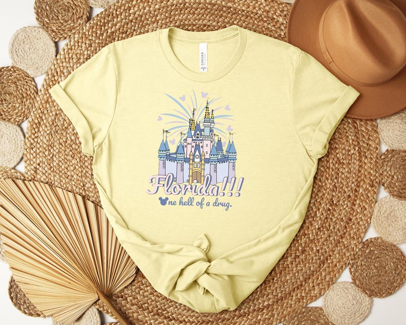 Puede incluir: Camiseta amarilla clara con una ilustraci&oacute;n colorida de un castillo. El texto dice "Florida!!! one hell of a drug." El dise&ntilde;o del castillo incluye detalles en azul, morado y dorado, con fuegos artificiales y corazones.