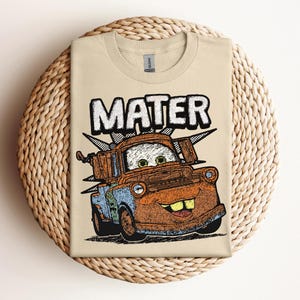 May include: A tan t-shirt featuring a distressed graphic of the character Mater from the movie Cars. The word "MATER" is printed above the character in a bold, outlined font. The graphic is in shades of orange, blue, and brown.
