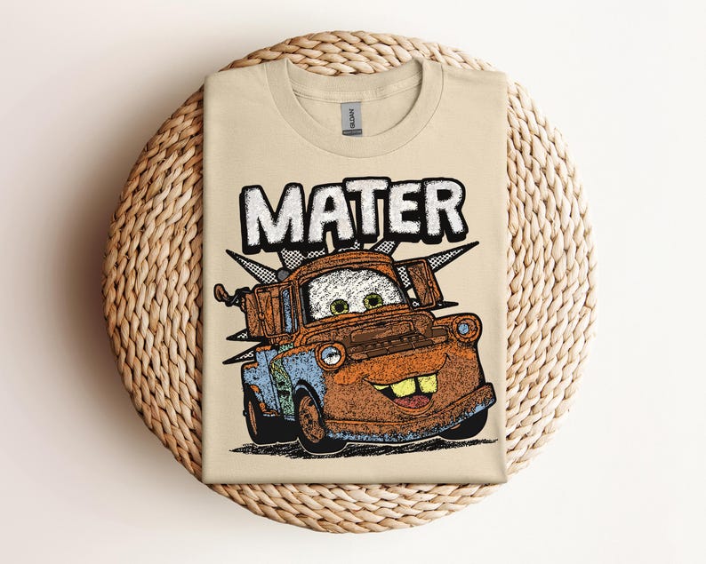 May include: A tan t-shirt featuring a distressed graphic of the character Mater from the movie Cars. The word "MATER" is printed above the character in a bold, outlined font. The graphic is in shades of orange, blue, and brown.