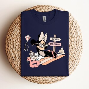 K&ouml;nnte beinhalten: Ein marineblaues T-Shirt mit einem Cartoon-Bild von Minnie Mouse, die eine Sonnenbrille und eine rosa Schleife tr&auml;gt. Sie liegt auf einem Strandtuch mit einem rosa aufblasbaren Flamingo, einer Flasche Sonnencreme und einem Schild mit der Aufschrift "Sunset" und "Ice-Cream".