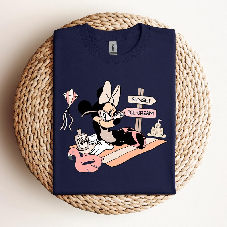 K&ouml;nnte beinhalten: Ein marineblaues T-Shirt mit einem Cartoon-Bild von Minnie Mouse, die eine Sonnenbrille und eine rosa Schleife tr&auml;gt. Sie liegt auf einem Strandtuch mit einem rosa aufblasbaren Flamingo, einer Flasche Sonnencreme und einem Schild mit der Aufschrift "Sunset" und "Ice-Cream".