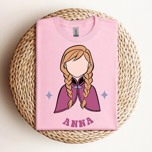 May include: A light pink t-shirt featuring a minimalist illustration of a character with blonde hair in two braids, wearing a magenta cape. The design includes two small blue four-pointed stars and the name ANNA printed in pink letters below.