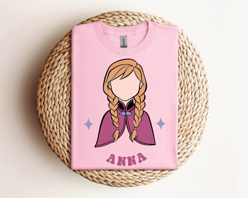 May include: A light pink t-shirt featuring a minimalist illustration of a character with blonde hair in two braids, wearing a magenta cape. The design includes two small blue four-pointed stars and the name ANNA printed in pink letters below.