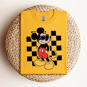 Pode incluir: Camiseta amarela com fundo xadrez preto e branco com o Mickey Mouse usando &oacute;culos de sol. O Mickey Mouse est&aacute; usando shorts vermelhos com um &uacute;nico bot&atilde;o preto.