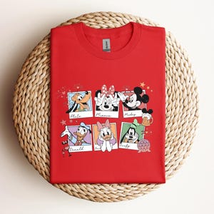 May include: A red short-sleeved t-shirt featuring a graphic print of six polaroid-style frames. Each frame contains a Disney character illustration with their name written below: Pluto, Minnie, Mickey, Donald, Daisy, and Goofy, along with small icons.