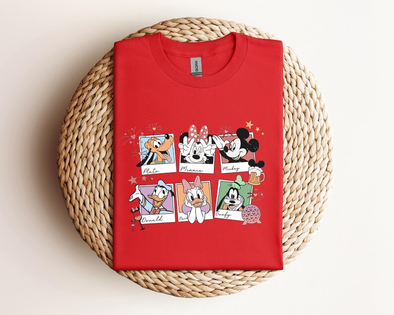 May include: A red short-sleeved t-shirt featuring a graphic print of six polaroid-style frames. Each frame contains a Disney character illustration with their name written below: Pluto, Minnie, Mickey, Donald, Daisy, and Goofy, along with small icons.
