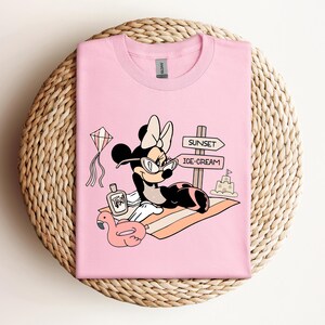 K&ouml;nnte beinhalten: Pinkes T-Shirt mit einem Cartoon-Bild von Minnie Mouse, die eine Sonnenbrille und einen Badeanzug tr&auml;gt und sich auf einem Strandtuch mit einem rosafarbenen Flamingo-Schwimmring, einer Flasche Sonnencreme und einem Schild mit der Aufschrift "Sunset" und "Ice-Cream" entspannt.