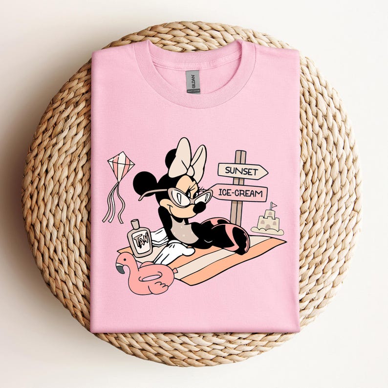 K&ouml;nnte beinhalten: Pinkes T-Shirt mit einem Cartoon-Bild von Minnie Mouse, die eine Sonnenbrille und einen Badeanzug tr&auml;gt und sich auf einem Strandtuch mit einem rosafarbenen Flamingo-Schwimmring, einer Flasche Sonnencreme und einem Schild mit der Aufschrift "Sunset" und "Ice-Cream" entspannt.