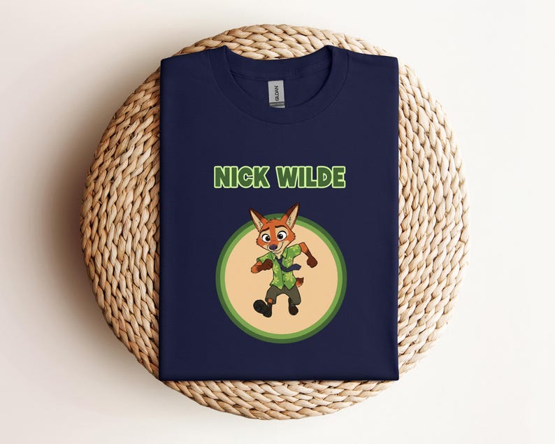 May include: Navy blue t-shirt featuring the text "NICK WILDE" in green above a cartoon image of a fox character. The fox is wearing a green shirt and brown pants, set against a light green and tan circular background.