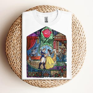 May include: White t-shirt featuring a colorful stained-glass design with characters from a fairy tale. The design includes a red rose, a couple dancing, and other characters. The shirt is folded on a woven, circular surface.