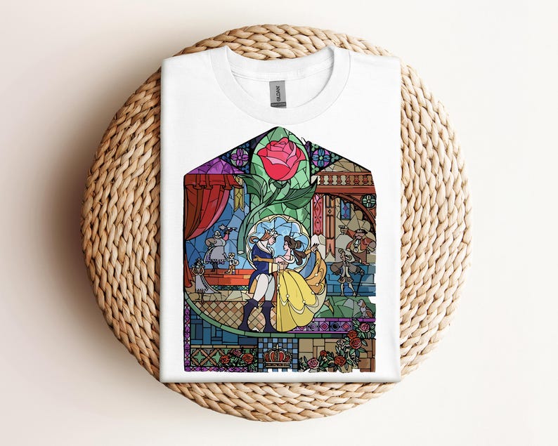 May include: White t-shirt featuring a colorful stained-glass design with characters from a fairy tale. The design includes a red rose, a couple dancing, and other characters. The shirt is folded on a woven, circular surface.