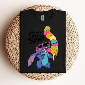 May include: Black t-shirt featuring a cartoon character wearing sunglasses and holding a colorful ice cream cone. The shirt has the words "hello summer" in a cursive font. The character is blue with pink accents.