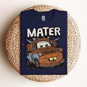 May include: Navy blue t-shirt featuring a graphic of Mater from the movie Cars. The word "MATER" is printed above the character in a white, distressed font. The cartoon Mater is brown, blue, and orange.