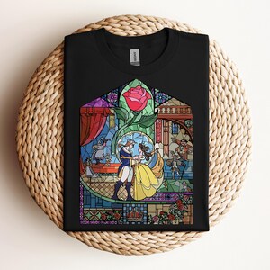 May include: Black t-shirt featuring a stained-glass design with characters from a fairy tale. The design includes a red rose, a couple dancing, and other characters. The shirt is folded on a woven surface.